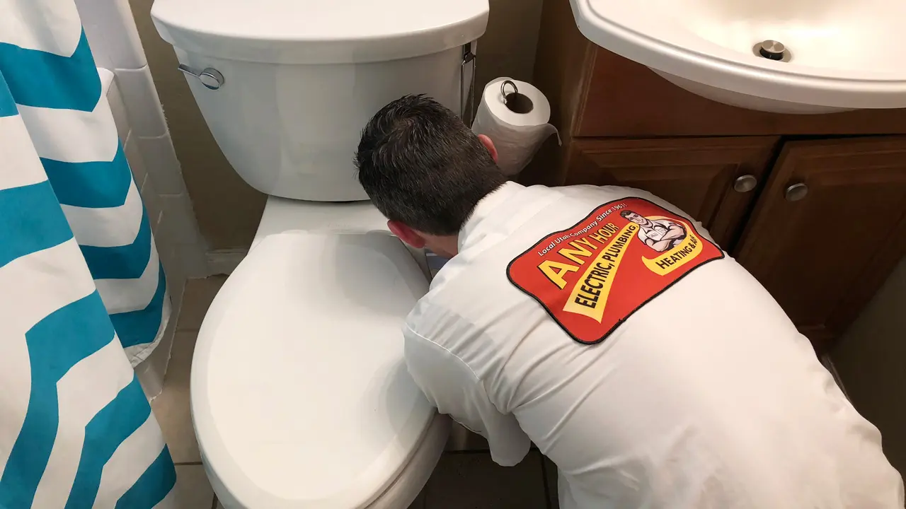 %Focuskw% | Any Hour Services A plumber in uniform kneels in front of a toilet, providing plumbing help and working on repairs to ensure the bathroom's plumbing system runs smoothly.