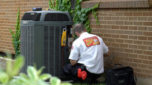 A technician in uniform checks and repairs an outdoor air conditioning unit next to a brick building, ensuring optimal room cooling for summer comfort.