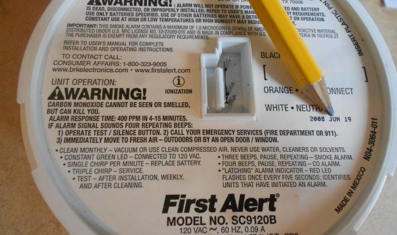 Close-up of a First Alert smoke and carbon monoxide detector with a yellow pencil pointing at the battery compartment. Warning and installation instructions highlight the serious business of preventing carbon monoxide poisoning.