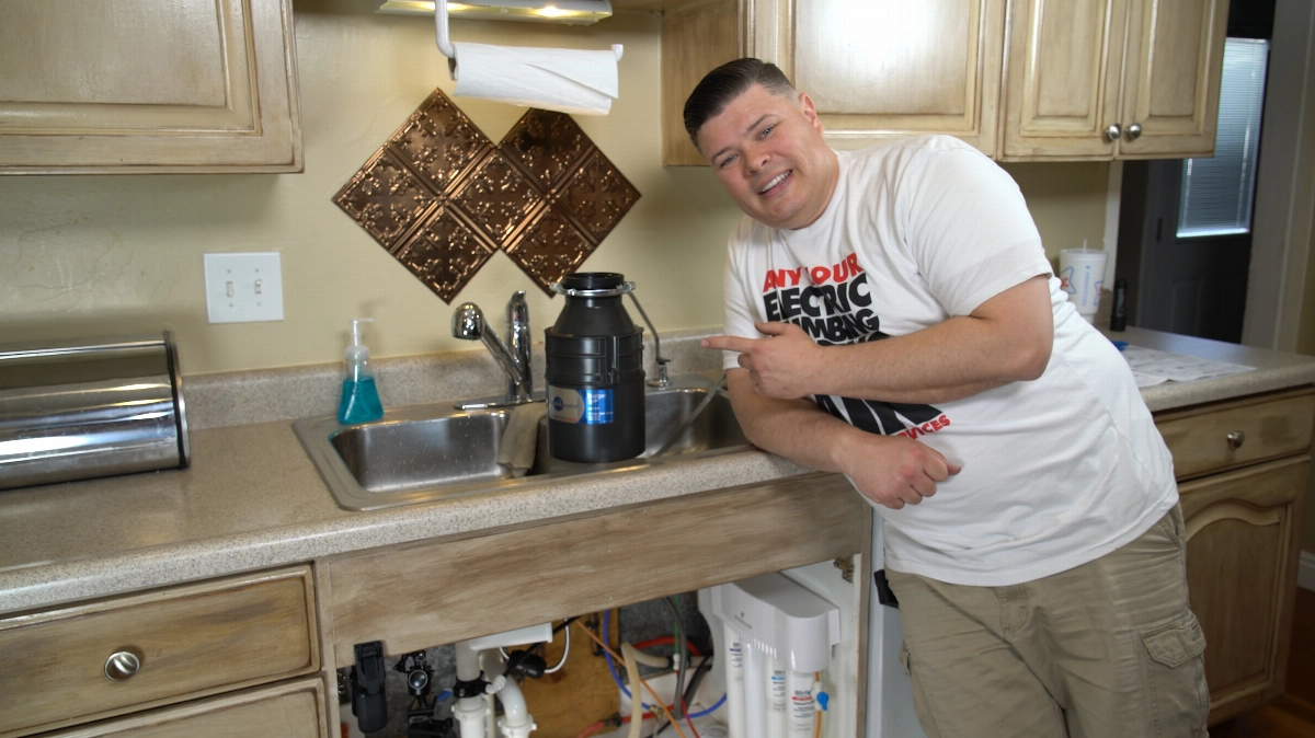 %Title% | Any Hour Services A man in a white t-shirt stands in a kitchen, smiling and pointing at a garbage disposal unit installed in the sink, ready to share maintenance tips for peak performance.
