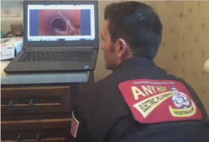 %Title% | Any Hour Services A technician in uniform examines a laptop displaying a pipe inspection image, checking for drain blockage possibly caused by tree roots, with the plumbing company branding visible on his jacket.