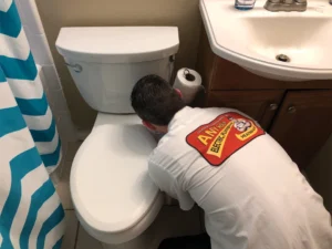 %Title% | Any Hour Services A plumber in a uniform is kneeling and inspecting or repairing a toilet next to a sink in a bathroom, demonstrating useful plumbing tips for homeowners.