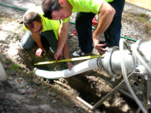Two workers in yellow shirts measure a white underground pipe, inspecting common sewer materials with a tape measure at an outdoor construction site.