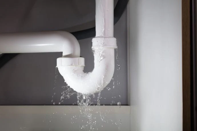 %Focuskw% | Any Hour Services Water leaking from a white plastic pipe under a sink, with visible water spraying from a crack in the curved section—an early sewer problem sign that could mean you need prompt sewer repair.