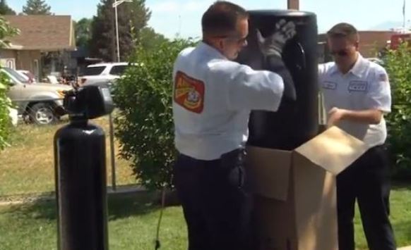 Two technicians in uniforms unpack and inspect a large black water softener tank outdoors near a cardboard box, with houses and greenery in the background, showcasing the water softener benefits for home use.