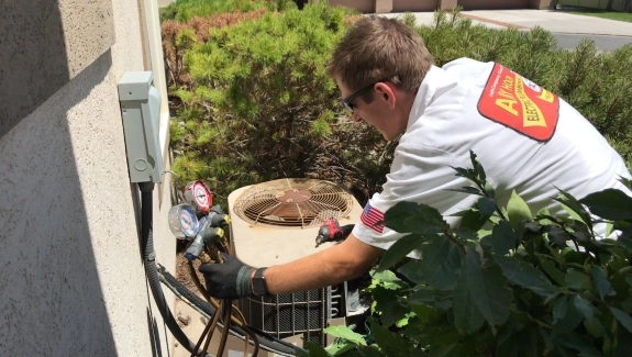A technician wearing a white shirt and gloves is performing professional AC maintenance on an outdoor air conditioning unit with gauges attached.