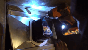 A person uses a power drill to install or remove screws from an HVAC system vent or duct component, with a sticker visible on the part.