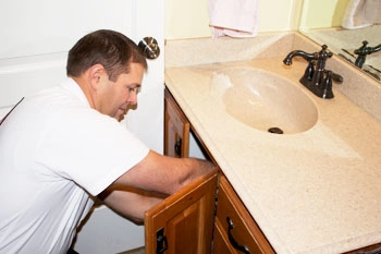 A man in a white shirt is fixing or inspecting plumbing under a bathroom sink with the cabinet door open.