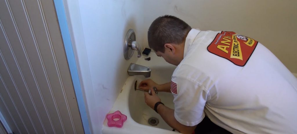 A plumber in uniform repairs the faucet of a white bathtub in a bathroom, helping address causes of sewer odors in the house. A pink bath toy is visible on the tub’s edge.