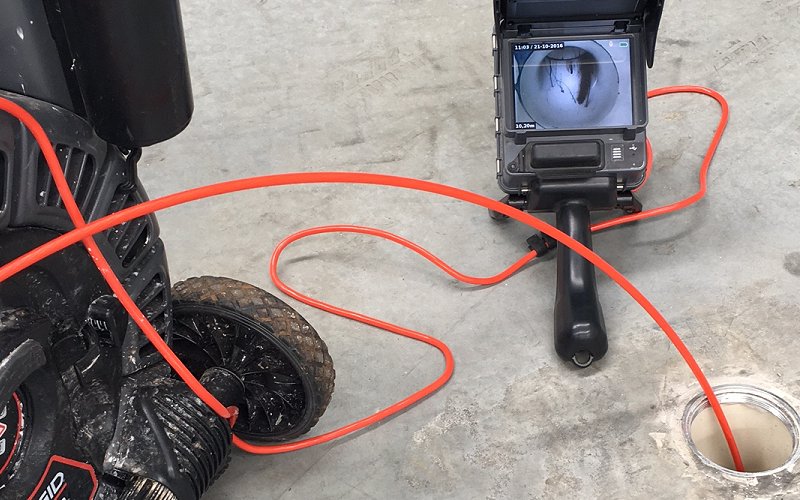 %Title% | Any Hour Services A sewer inspection camera with a red cable is positioned next to an open drain, its monitor displaying a potential drain blockage inside the pipe on a concrete floor.
