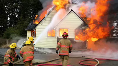 Firefighters spray water on a house engulfed in flames, with heavy smoke and fire coming from the roof and windows.