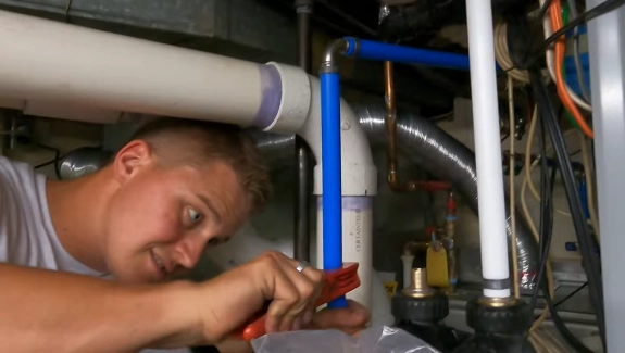 %Title% | Any Hour Services A person uses a wrench to tighten a blue PEX plumbing pipe connection under a sink amid various plumbing and electrical components.