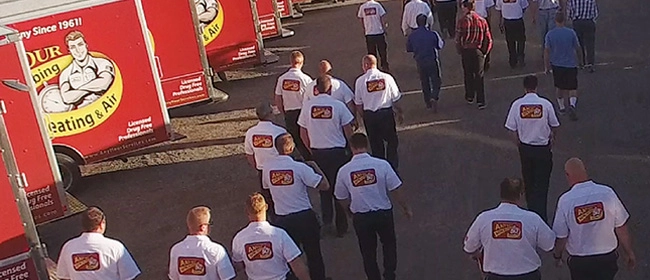 A group of men in white shirts with company logos walk towards red company trucks parked in a lot.