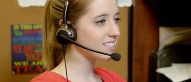 A woman wearing a headset and red shirt is smiling while working at a desk, with papers and a colorful sign in the background.