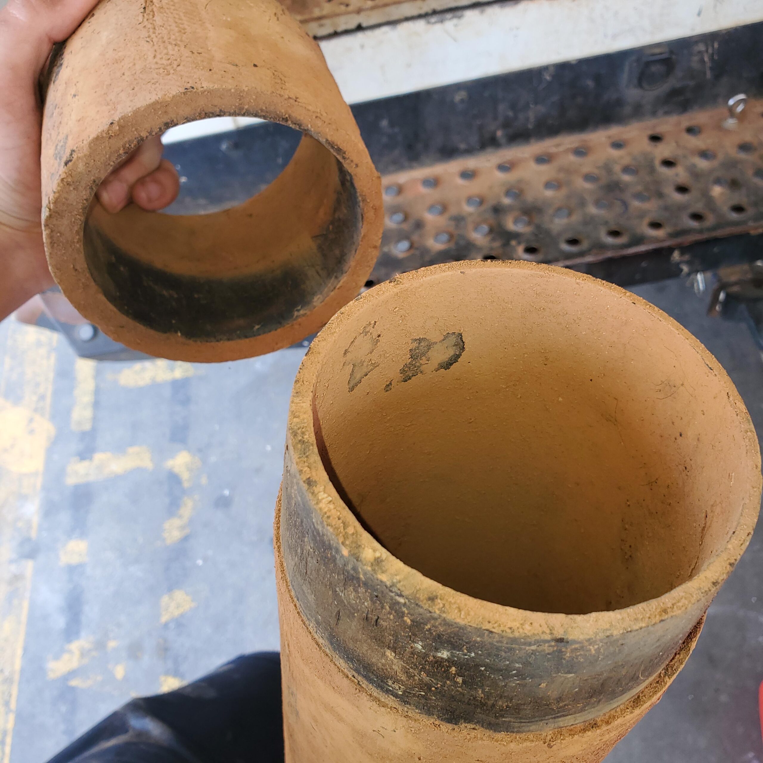 Two hands hold sections of a brown ceramic sewer pipe—one of the common sewer materials—showing black residue inside, positioned above a metal perforated surface.