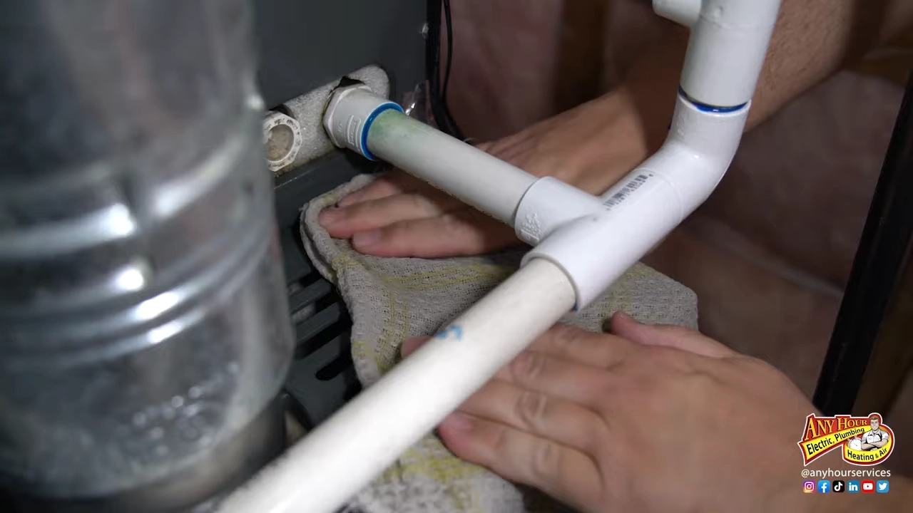 %Title% | Any Hour Services Close-up of hands using a cloth to clean around white PVC pipes connected to a metal unit, likely part of winter furnace maintenance for a home HVAC system. Any Hour Services logo in the bottom right corner.