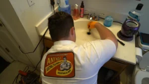 %Title% | Any Hour Services A plumber wearing a company shirt expertly unclogs a bathroom sink drain with tools, demonstrating essential plumbing maintenance; various cleaning items and toiletries are on the sink.