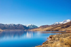 %Title% | Any Hour Services A clear blue lake reflects nearby snow-capped mountains and hills under a cloudless sky, with dry grass along the shoreline—an area where rising water tables could increase flooding risk in the future.