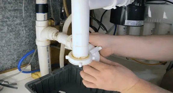 %Imagetitle% | Any Hour Services Hands working on PVC plumbing pipes under a sink, likely performing kitchen sink drain cleaning or repair, with a black tray placed underneath to catch any water.