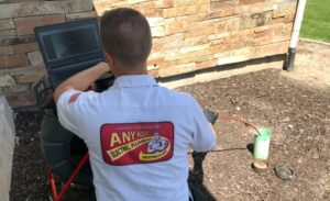 %Title% | Any Hour Services A technician in an "Any Hour Electric, Plumbing, Heating & Air" shirt inspects outdoor plumbing with a laptop and equipment near a stone wall, checking for sewer system problems and potential sewer main failure.