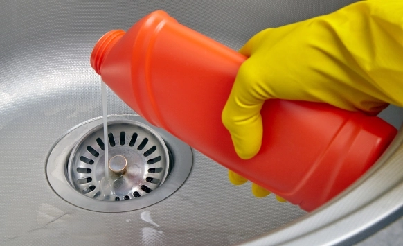 A person wearing yellow gloves pours liquid chemical drain cleaner from a red bottle into a metal sink drain, highlighting common chemical drain cleaner problems during routine drain cleaning.