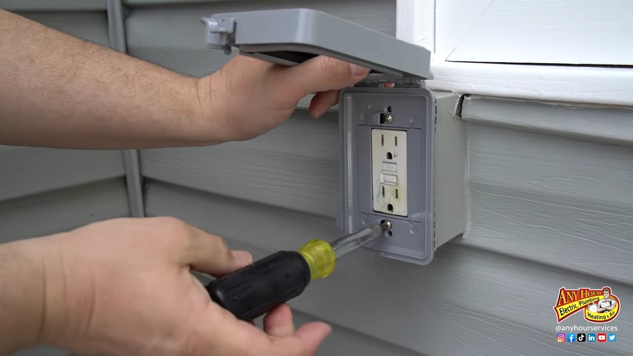 A person uses a screwdriver to install or repair an outdoor electrical outlet box on a house’s exterior wall, helping ensure Halloween fire safety when setting up seasonal lights and decorations.