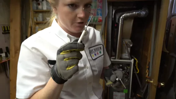 A technician wearing gloves holds a furnace ignitor, standing next to an open furnace in a utility room.