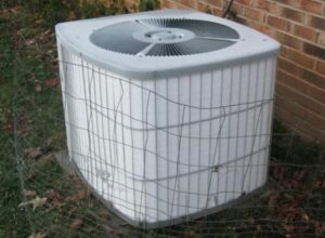 Outdoor air conditioning unit enclosed by a wire fence, situated next to a brick wall with some grass and leaves on the ground—a common spot where AC troubleshooting may be needed if your AC freezes over.