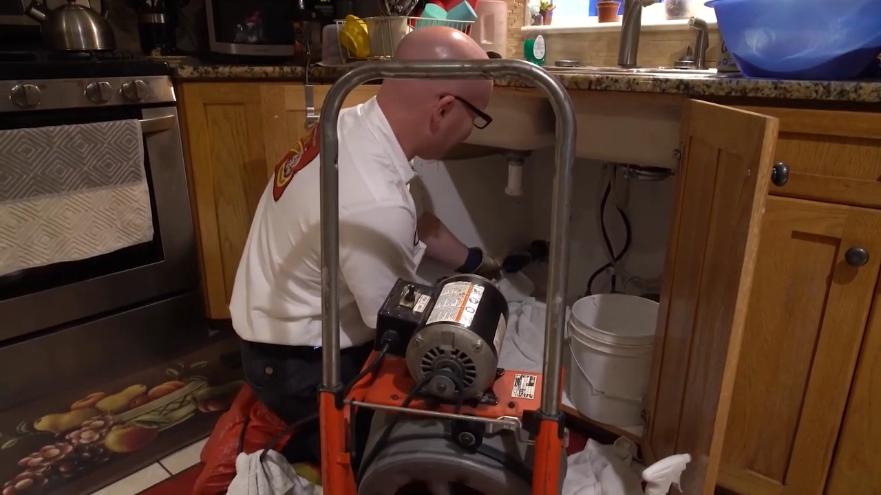 %Title% | Any Hour Services A plumber wearing gloves works under a kitchen sink, using a drain cleaning machine with a bucket nearby—demonstrating the importance of yearly drain cleaning for proper drain maintenance.