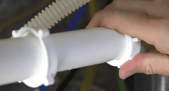%Imagetitle% | Any Hour Services A hand is tightening a white plastic plumbing pipe fitting under a clean kitchen sink drain, with a flexible hose and colored wires visible in the background.