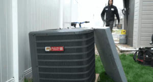 %Title% | Any Hour Services An outdoor AC unit with its cover removed sits on grass near a fence, while a person in work attire approaches in the background.