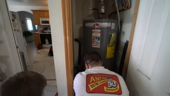 A technician in an "Any Hour Electric, Plumbing, Heating & Air" shirt works on a water heater in a small utility closet near a kitchen.