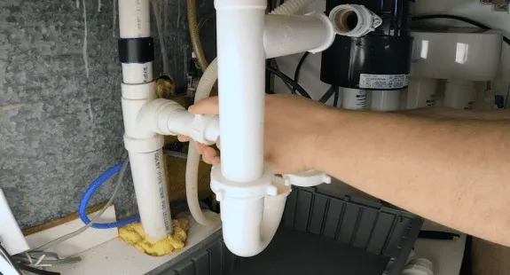 %Imagetitle% | Any Hour Services A person’s hand is holding PVC pipes under a sink, appearing to perform plumbing work—perhaps learning how to clean sink drain or fix a smelly sink drain issue near the drain assembly.