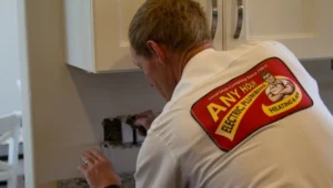 %Title% | Any Hour Services A technician wearing an "Any Hour Electric, Plumbing, Heating & Air" shirt works on an electrical outlet in a kitchen.
