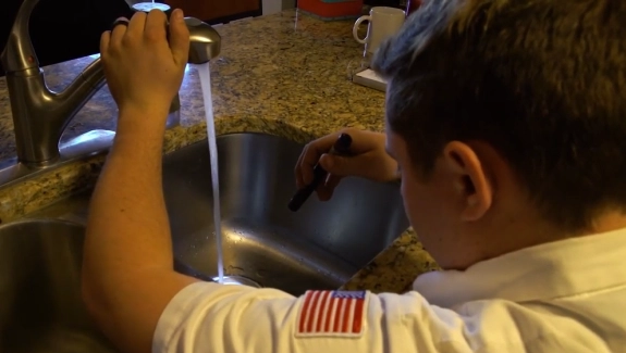 A person in a white shirt with a U.S. flag patch inspects a running kitchen faucet with a flashlight over a granite countertop.