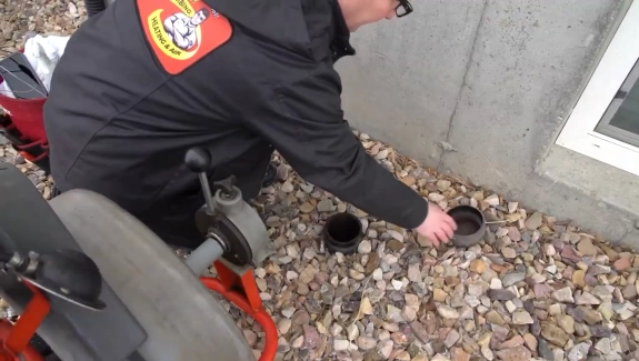 Person in black jacket kneeling on rocks, removing the cap from an outdoor drain cleanout pipe next to a building foundation—a common step in home wastewater or septic system maintenance. A drain cleaning machine is nearby.