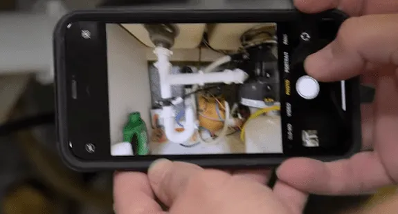 %Imagetitle% | Any Hour Services A person takes a photo of plumbing pipes under a sink with a smartphone, documenting the steps for how to clean sink drain and keep a clean kitchen sink drain.