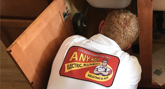 A technician wearing an "Any Hour Electric, Plumbing, Heating & Air" uniform works under a sink with cabinet doors open, ready to tackle your kitchen plumbing or answer any plumbing questions you have.