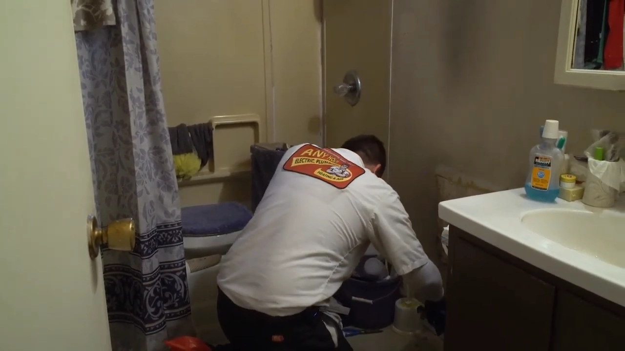 %Title% | Any Hour Services A plumber in uniform kneels on the bathroom floor, working near the toilet and bathtub with tools and cleaning supplies around him, ready to tackle common plumbing myths with expertise.