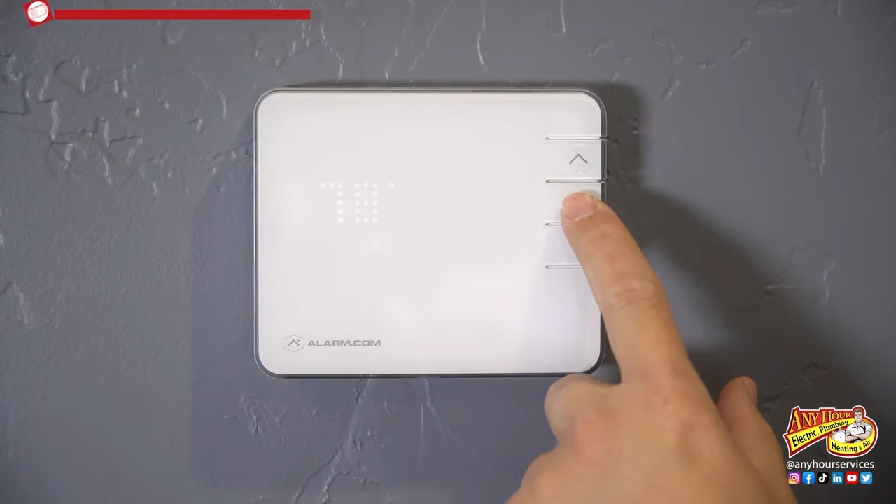 %Title% | Any Hour Services A hand presses the up arrow button on a white Alarm.com thermostat mounted on a gray textured wall, ensuring the furnace keeps your home cozy for winter.