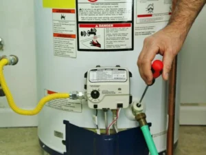 A person performs maintenance on a water heater, using a screwdriver to adjust or repair the control panel with warning labels visible, helping to extend its lifespan.