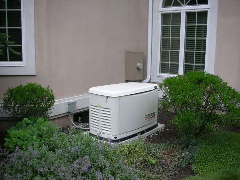 An expertly installed standby generator from Any Hour Services is positioned on a concrete pad beside a home in St. George, UT. Surrounded by lush bushes and plants near a window, this setup highlights how our team integrates home generator solutions smoothly into your landscape for both reliability and curb appeal.