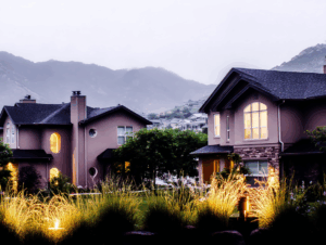 Two modern suburban houses in Utah feature illuminated landscaping, trees, and tall grasses in the front yard, set against misty mountains—perfect for enjoying safe furnace operation and carbon monoxide safety during colder seasons.