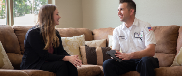 A woman and a technician sit on a living room couch, engaged in conversation. The technician wears a uniform and holds a tablet.